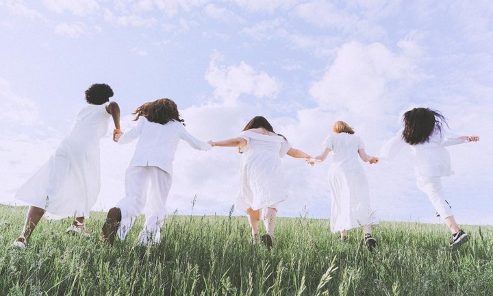 cinco mulheres vestidas de branco de mãos dadas correndo num campo